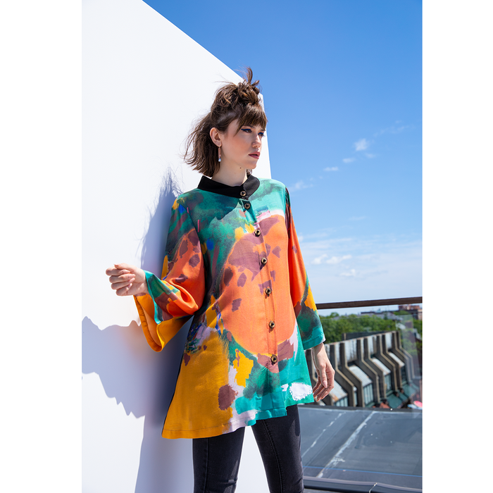 A model wearing a colorful abstract print top with 3/4 sleeves and a button-up front, standing against a plain background.