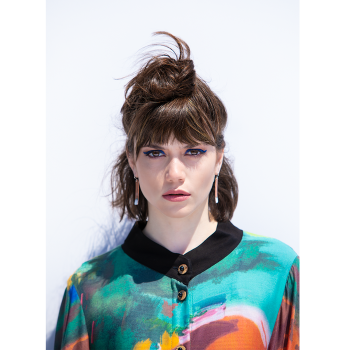 A model wearing a colorful abstract print top with 3/4 sleeves and a button-up front, standing against a plain background.