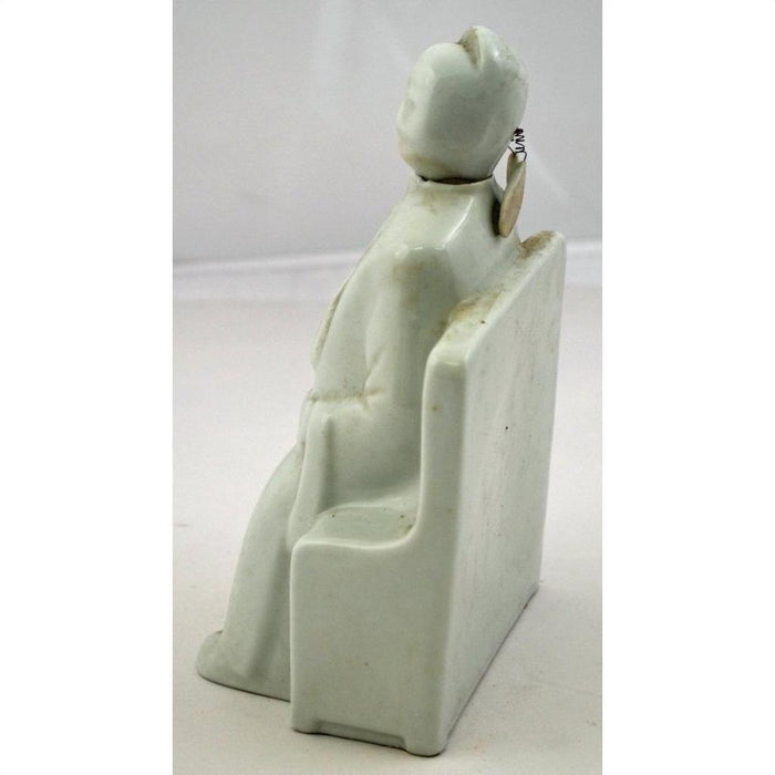 White Blanc de Chine porcelain doll with bobbing head, seated on bench, coin attached to head, close-up view.