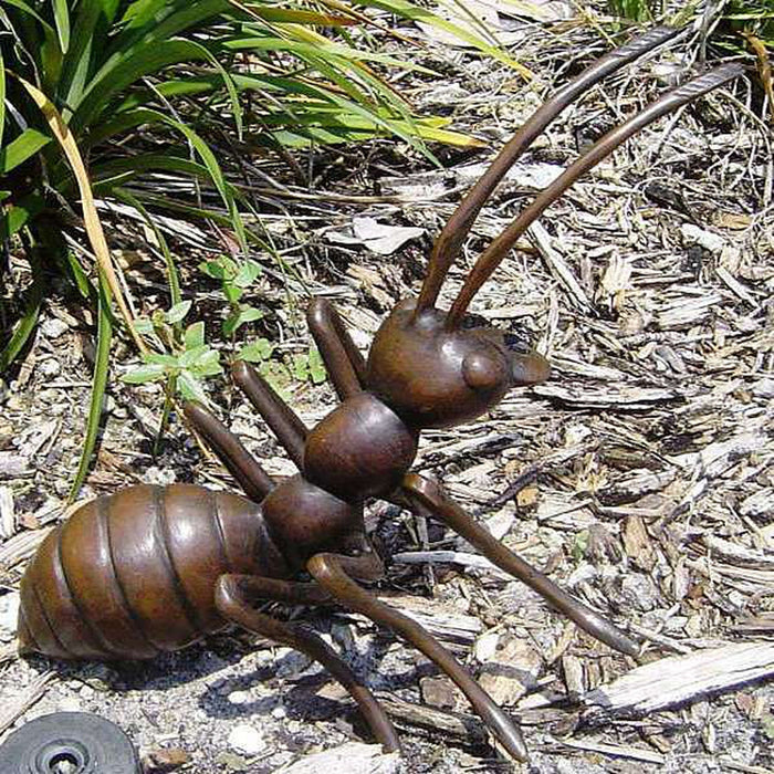 Bronze metal ant statue on garden mulch surrounded by green plants and grass, outdoor setting.