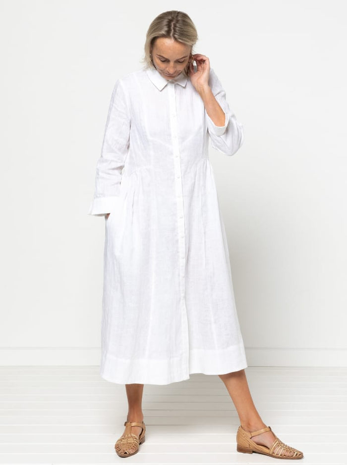 Woman wearing a white dress made of Hampton Woven Dress Sewing Pattern – Mid-calf shirtdress with gathered skirt, curved waist, sleeve or sleeveless options, and inseam pockets. against a plain background