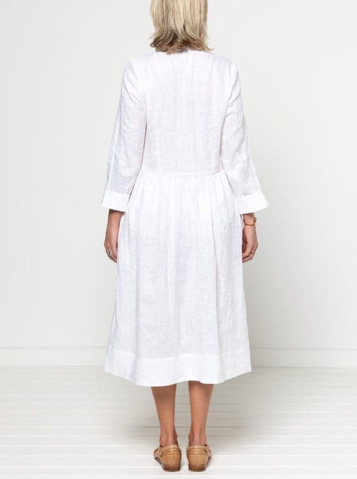 Person wearing a white dress against a plain background -Hampton Woven Dress Sewing Pattern – Mid-calf shirtdress with gathered skirt, curved waist, sleeve or sleeveless options, and inseam pockets.