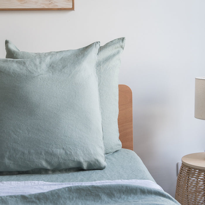 Harbour Sage European linen pillowcases  on a bed with a wooden headboard and lamp in the background.