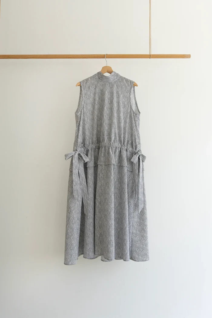 A sleeveless gray and white textured dress with side tie details, hanging on a wooden hanger from a wooden clothing rack against a plain white wall.