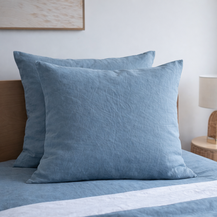 Two Lake Blue 100% Linen European Pillowcase on a bed with a lamp and wooden headboard in the background.