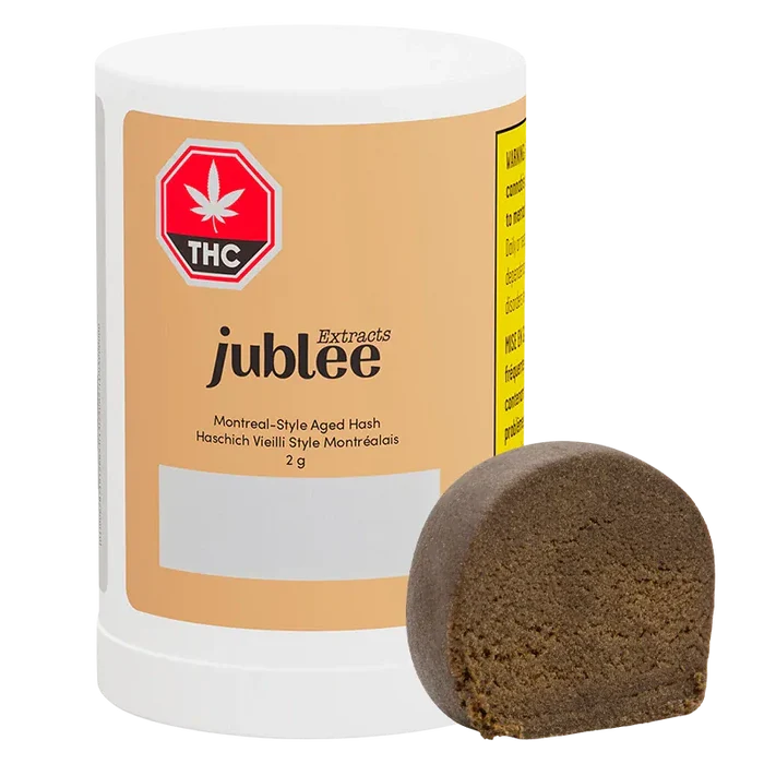 Front-facing shot of the Jubilee Extracts Montreal-Style Aged Hash 2 g cylinder with a brown half-disc of hash leaning against it.