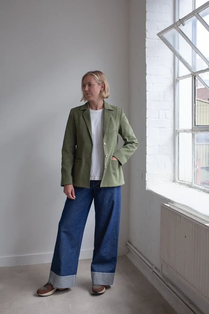 Woman wearing a green jacket made using Naomi Blazer PDF Sewing Pattern  and blue jeans standing in a room with a window.