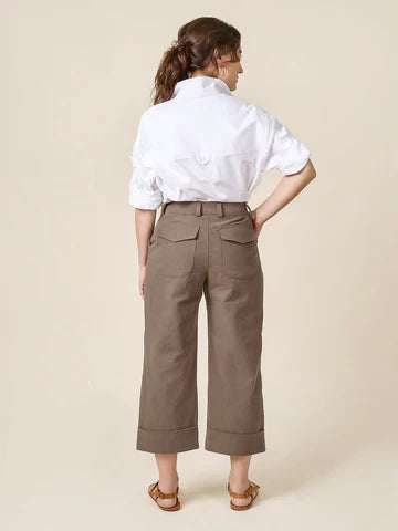 Woman wearing a white shirt and beige pants made of Rika Pants sewing pattern in cropped tailored style with hem cuffs and tabbed waistband against a beige background