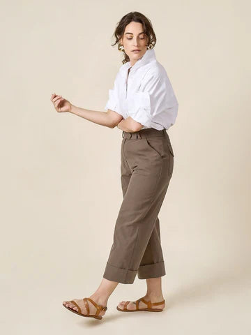 Woman wearing a white shirt and brown pants made of Rika Pants sewing pattern in cropped tailored style with hem cuffs and tabbed waistband  on a beige background