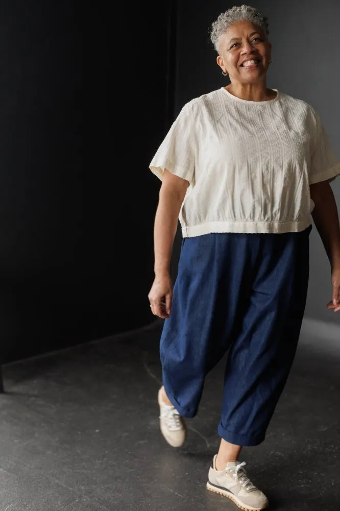 Woman wearing a white top top made using The Celeste Sewing Pattern by Merchant & Mills and blue pants against a black background