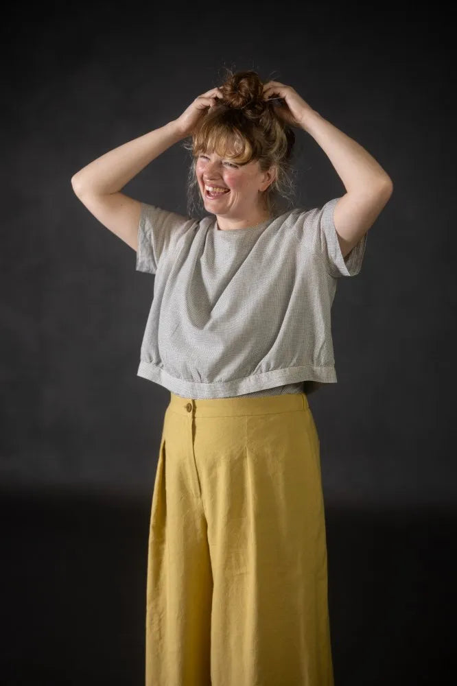 Person wearing a light grey top made using The Celeste Sewing Pattern by Merchant & Mills, and mustard yellow pants against a dark background