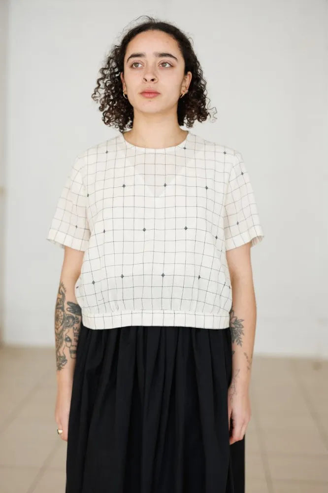 Person wearing a white checkered top made using The Celeste Sewing Pattern by Merchant & Millsand black pants against a plain background