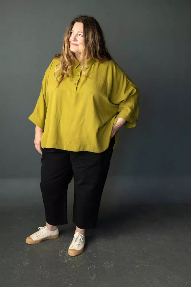Woman wearing a mustard yellow blouse and black pants  made using The Eve Trouser Sewing Pattern by Merchant & Mills – cropped tapered trousers with side zip and pleats against a gray background