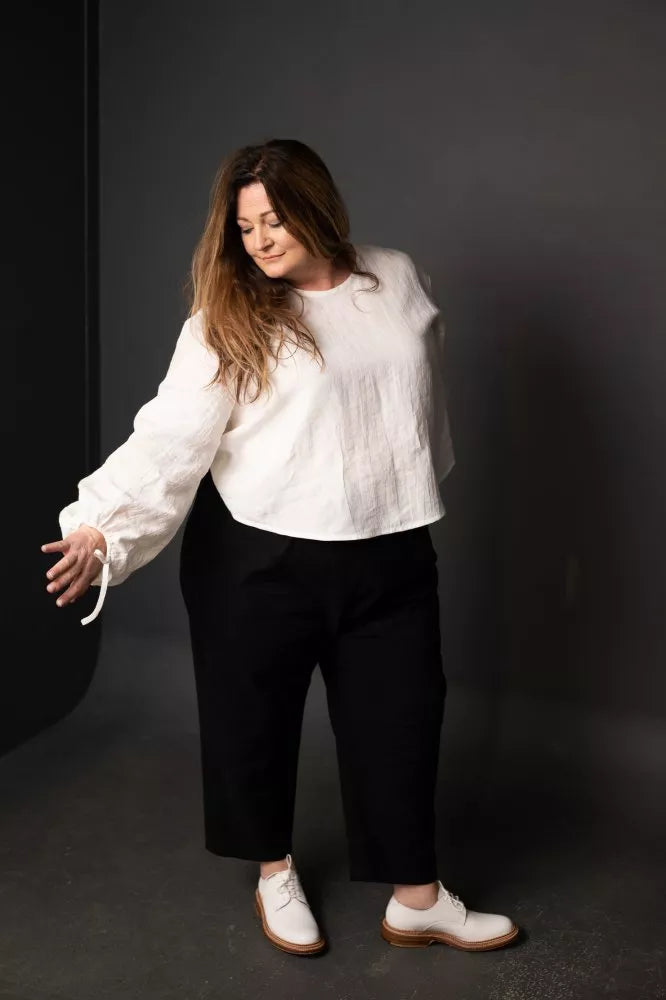 Person wearing a white blouse and black pants made using The Eve Trouser Sewing Pattern by Merchant & Mills – cropped tapered trousers with side zip and pleats on a dark background