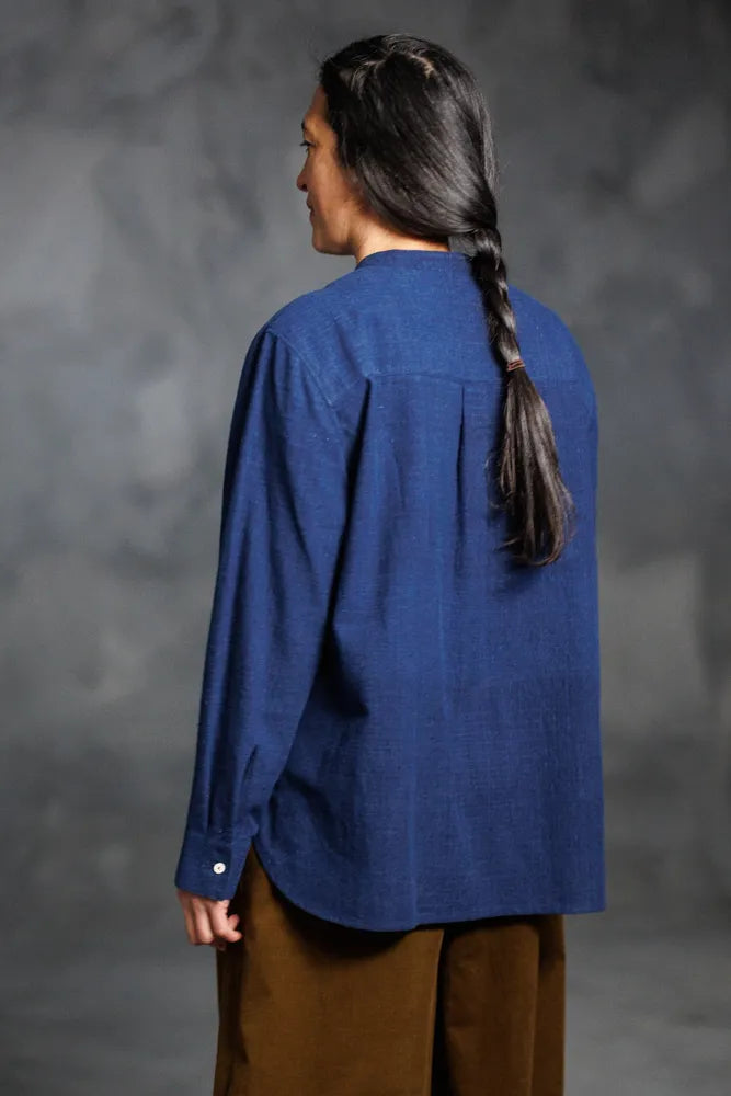 Person wearing a blue top made using The Niven Sewing Pattern by Merchant & Mills – oversized shirt or shirt-dress with bib, grandad collar and cuffs and brown pants against a gray background