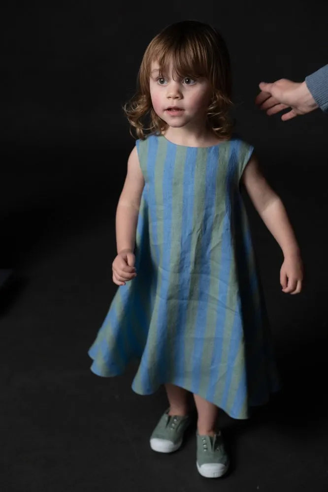 Young girl wearing a blue and green striped dress  made using trapezette girls dress sewing pattern against a dark background