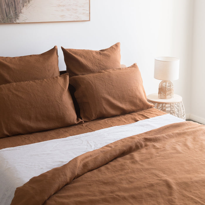 Tobacco brown linen fitted sheet on a bed with a lamp and wicker side table in the background.